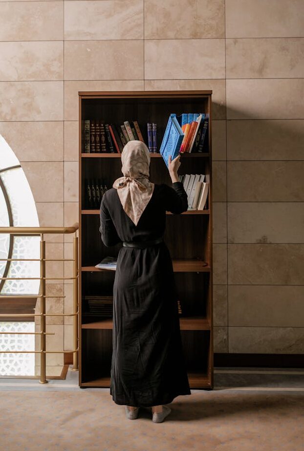 Muslim woman in hijab choosing books from a bookshelf indoors.